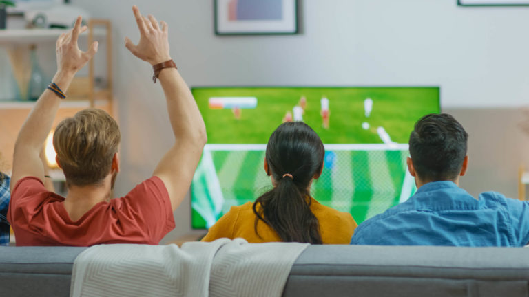 Three friends sitting on couch watching soccer game while one friend is cheering