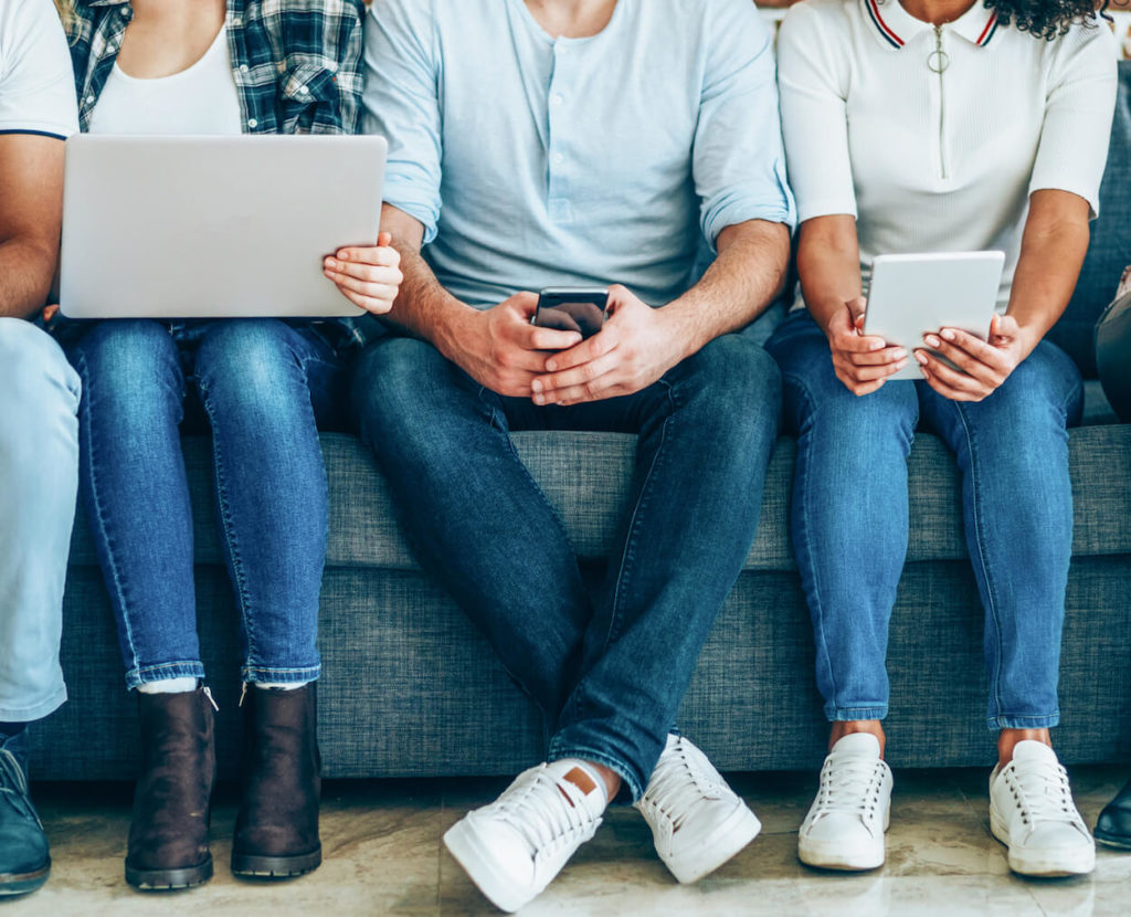 A group of young men and women hold internet-enabled devices while sitting on a couch