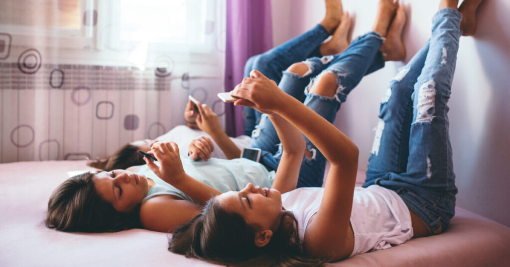 Three teenage girls laying on a bed with their feet up on the wall while looking at their smartphones.