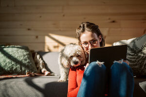 A woman sits on a couch with her poodle dog and uses her tablet