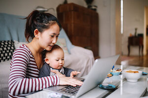 A young Asian woman works on her laptop with her infant boy in her lap
