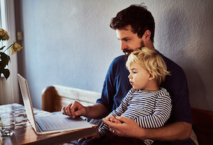 A man sits with a young boy on his lap while using a laptop