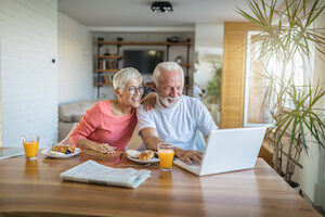 An elderly couple sits at the dining table and uses a laptop