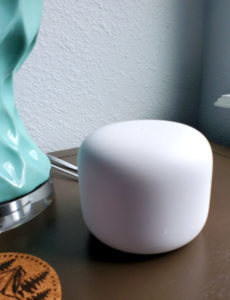 A Google Nest Wi-Fi router sits on a table next to a blue lamp