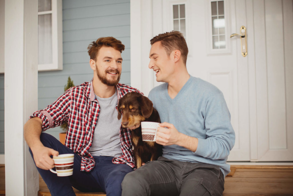 Young couple sitting with their dog on front porch of house, holding mugs
