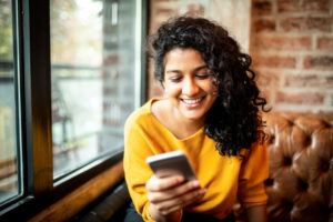 A woman with curly, dark hair and a yellow shirt looks at her phone and smiles