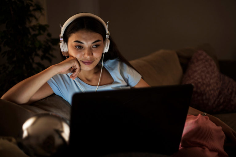 Woman watching NBC Peacock on laptop sitting on couch in the dark with headphones on