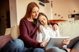 A woman and her daughter play on a tablet while sitting on the couch together