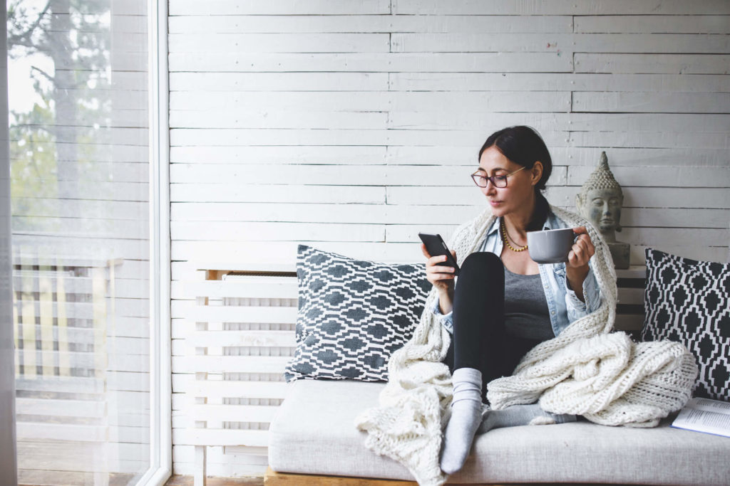 Woman sitting on a couch looking at her phone and holding a mug