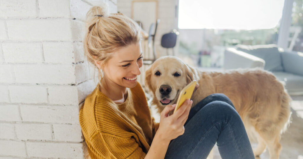 A woman is sitting on the ground with her Labrador while looking at her smartphone.