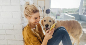 A woman is sitting on the ground with her Labrador while looking at her smartphone.