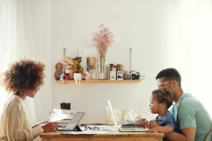 Una mamma, papà e figlio afroamericani si siedono alle estremità opposte di un tavolo mentre lavora online