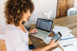 A woman works on her laptop and looks at her cell phone.