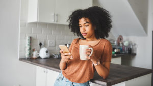 A woman looks at her phone while drinking coffee in the kitchen.