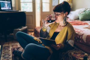 Woman sitting on floor in bedroom watching Hallmark Channel on tablet while drinking wine