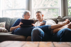 An older White man and woman lounge on a couch with their tablet and cell phone