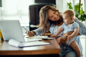 A woman holds her infant in her lap while she works on her laptop