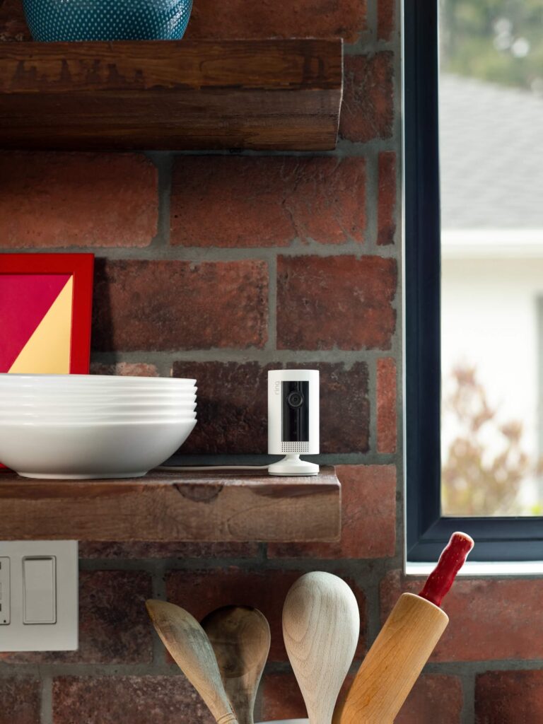 Ring Indoor Cam on wooden shelf above some kitchen utensils and next to a stack of bowls