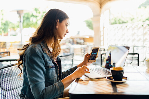 A brunette woman looks at her cell phone and laptop screen