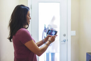 Woman looks at live view of video doorbell on her phone. In the background, you can see the outline of a visitor through the door.