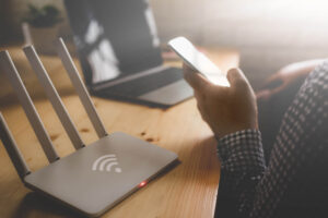 A router and a laptop sit on a desk. A hand is hold a cellphone, but the rest of the person is off frame