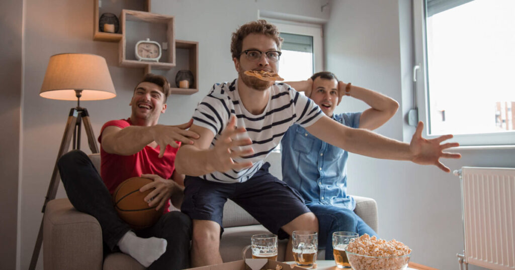 Group of friends watching basketball in living room while drinking beer and eating pizza and popcorn