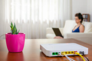 An internet router sits in the foreground while a woman uses a laptop in the background