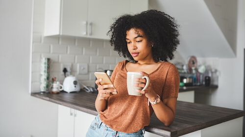 A young Black woman leans against the counter while she looks at her cellphone