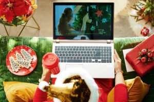 Woman streaming Christmas movie on laptop while sitting on couch, eating cookies, and drinking hot cocoa