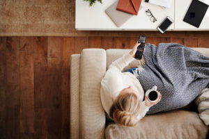 Overhead Shot Looking Down On Woman At Home Lying On Sofa, Drinking Coffe, and Watching TV