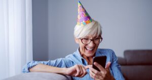 Woman with glasses and small birthday hat smiling while using her smartphone