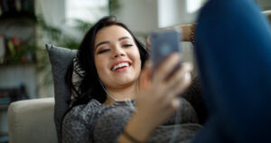 Young woman is wearing headphones and smiling while looking at her iPhone.