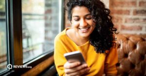 A woman with curly, dark hair and a yellow shirt looks at her phone and smiles