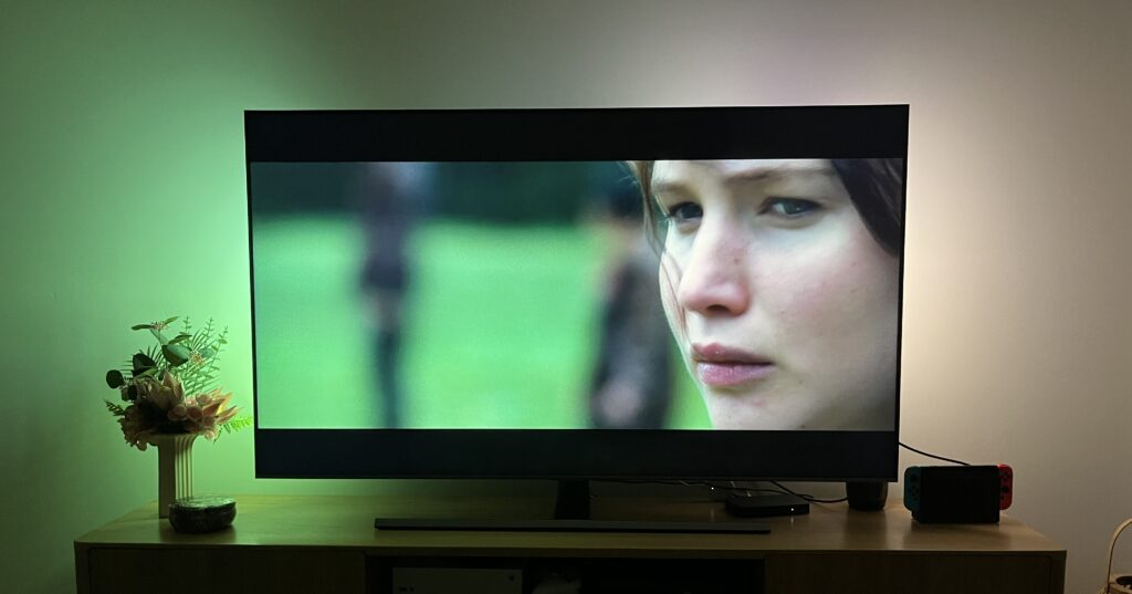 The Hunger Games playing on a 65-inch TV. Behind the TV, the Philips Hue Play Gradient Lightstrip is reflecting the colours playing on the screen.