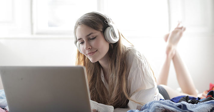 young woman lying on a bed wearing headphones and watching a movie on her laptop