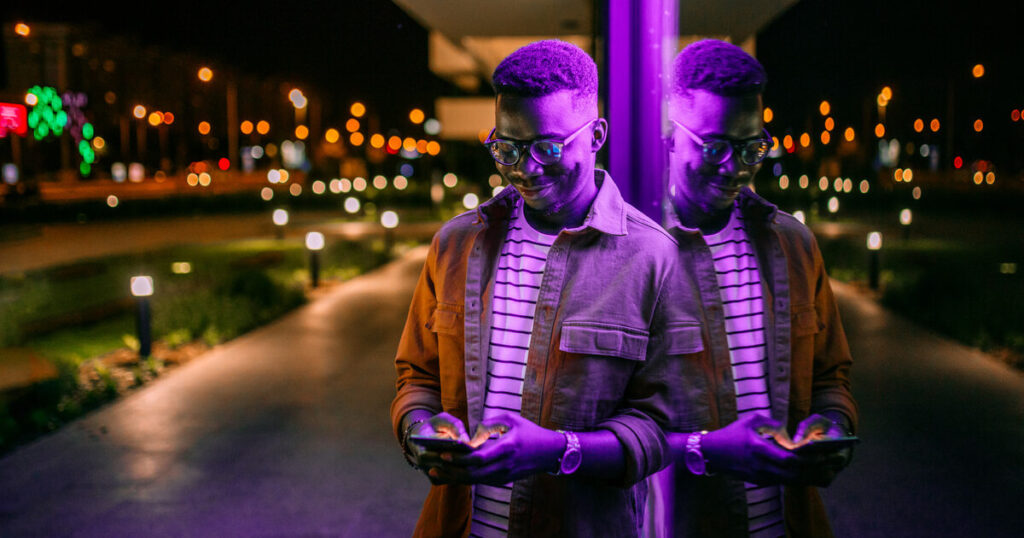 Man leans against glass bus stop while looking down at this phone