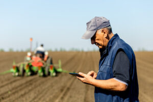 An older farmer looks at his tablet while another farmer plows a field in the background