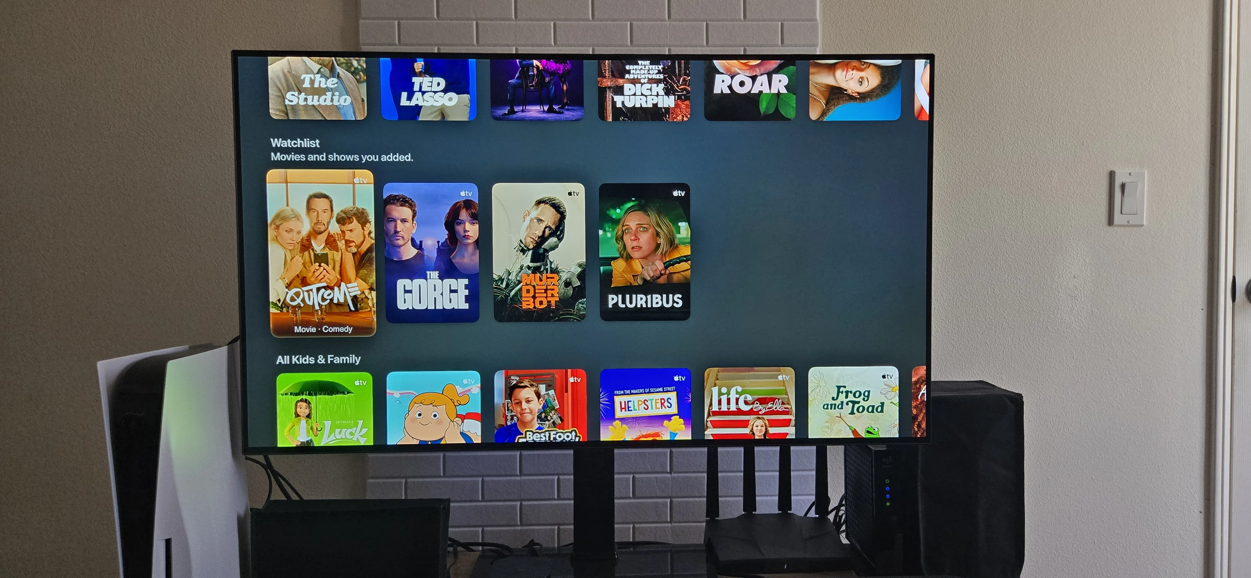 Apple TV+ streaming interface displayed on a large TV screen showing the Watchlist section with titles including The Gorge, Murderbot, and Pluribus, along with Kids & Family content