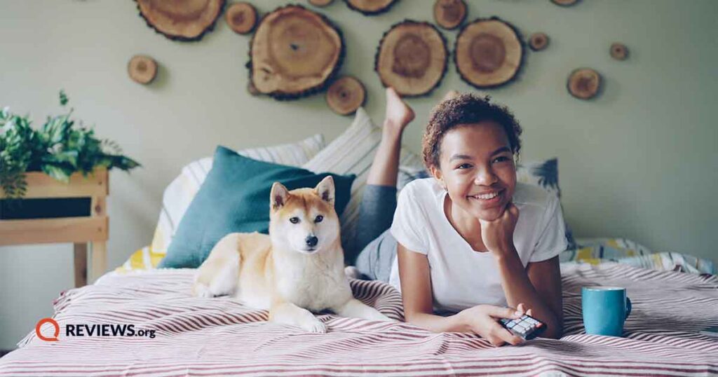 happy black female laying on bed with remote watching tv and sitting next to her dog