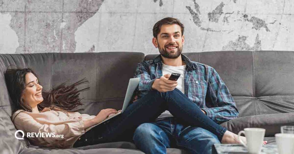 male and female sitting on a gray couch while one watches tv and one uses a laptop