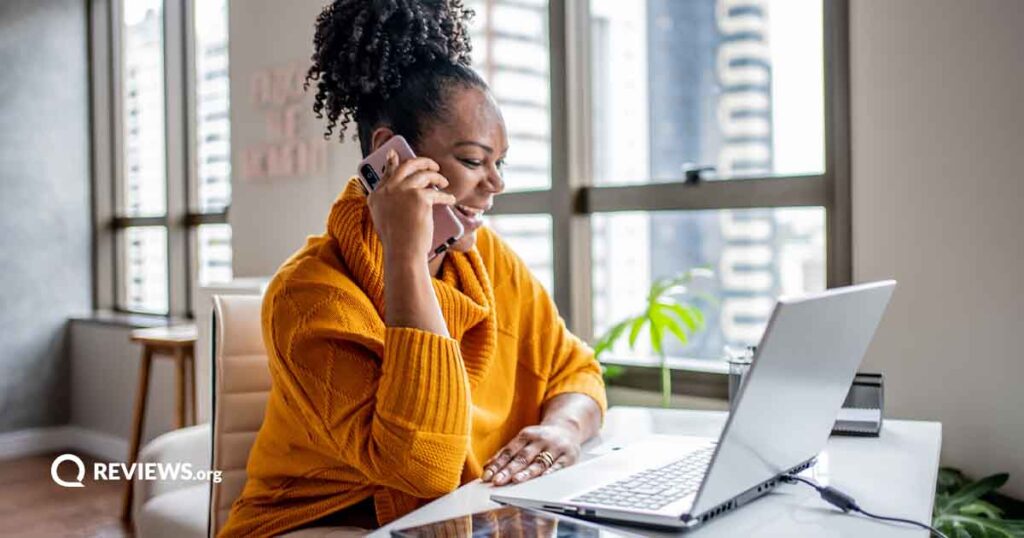 happy black woman sitting at desk while talking on the phone and using a laptop