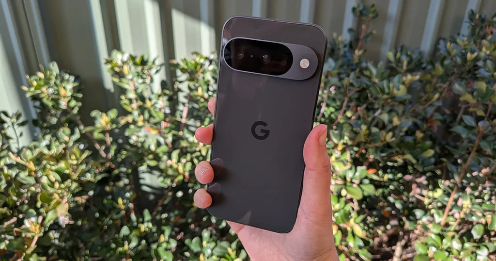 A hand holding an Obsidian Google Pixel 10 against a background of green bushes