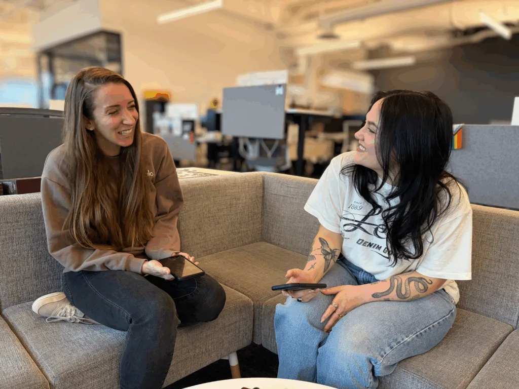 Two coworkers sit on a gray couch in an office lounge, smiling at each other while holding phones.