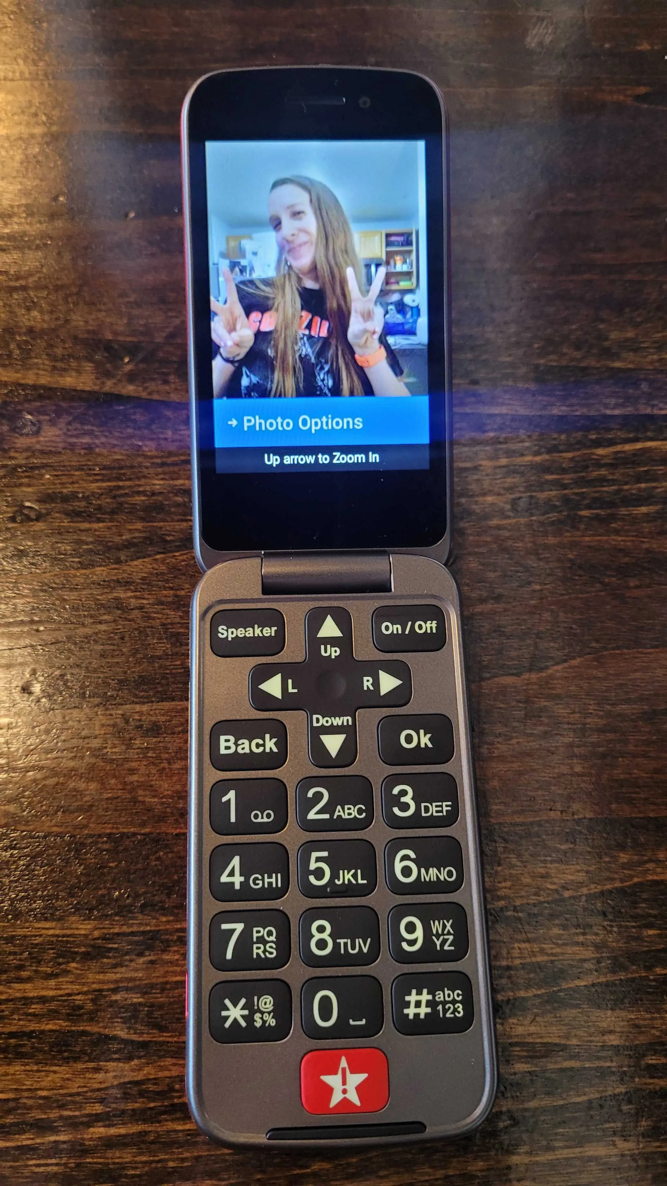 A Lively Jitterbug Flip2 phone shown open on a wooden surface. The top screen displays a photo of a person making peace signs, while the bottom half features an oversized, backlit physical keypad with high-contrast white numbers on black buttons. A prominent red "Urgent Response" button with a star and exclamation point icon is located at the very bottom center.