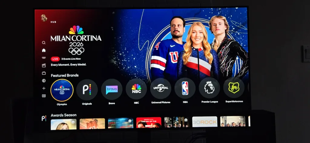 A smart TV displaying the Peacock homepage featuring a large banner for the Milan Cortina 2026 Winter Olympics. Below the banner, a "Featured Brands" row includes icons for the Olympics, Peacock Originals, Bravo, NBC, Universal Pictures, NBA, Premier League, and SuperMotocross