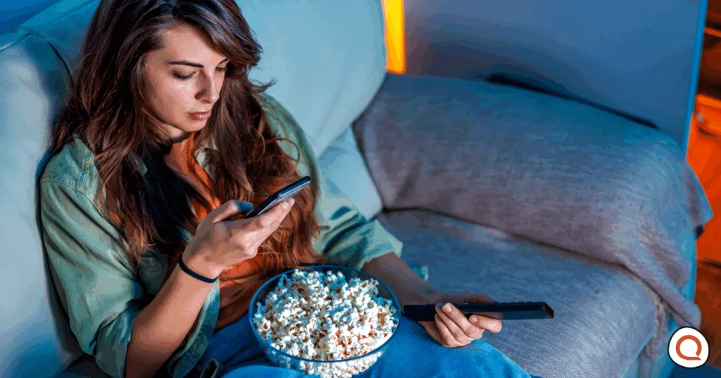 Woman sitting on couch with remote and popcorn looking at her phone. Image credit: iStock/vladans