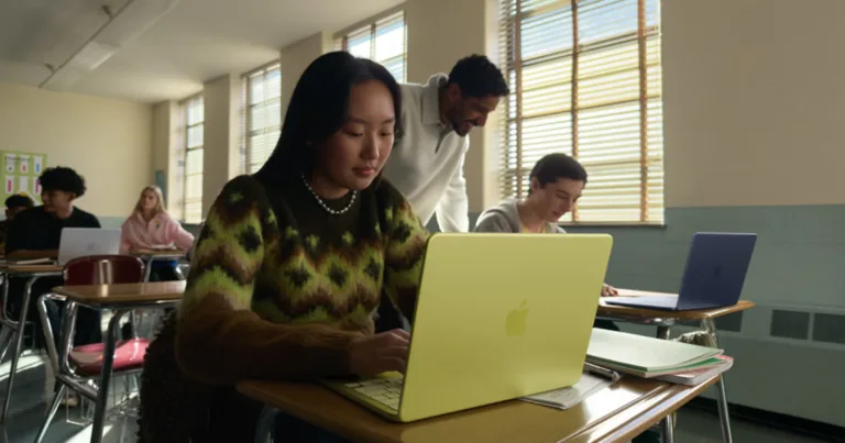 High school students using Apple MacBooks in a classroom while a teacher assists at their desks