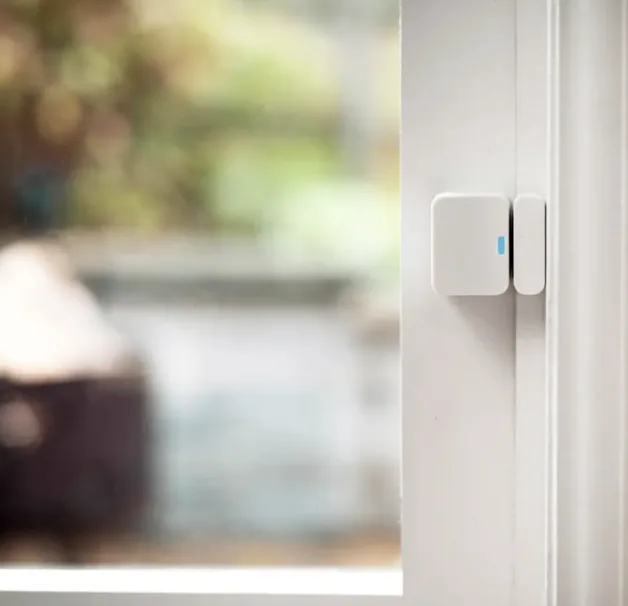 A SimpliSafe wireless entry sensor mounted on a white window frame, with a blurred outdoor view through the glass in the background.