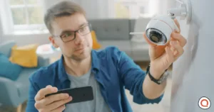 Man installing a wireless outdoor home security camera while monitoring setup on smartphone for burglary protection