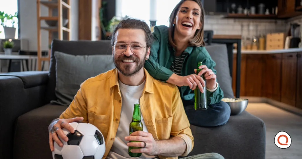 Couple watching soccer at home on the couch, smiling and holding beers and a soccer ball.